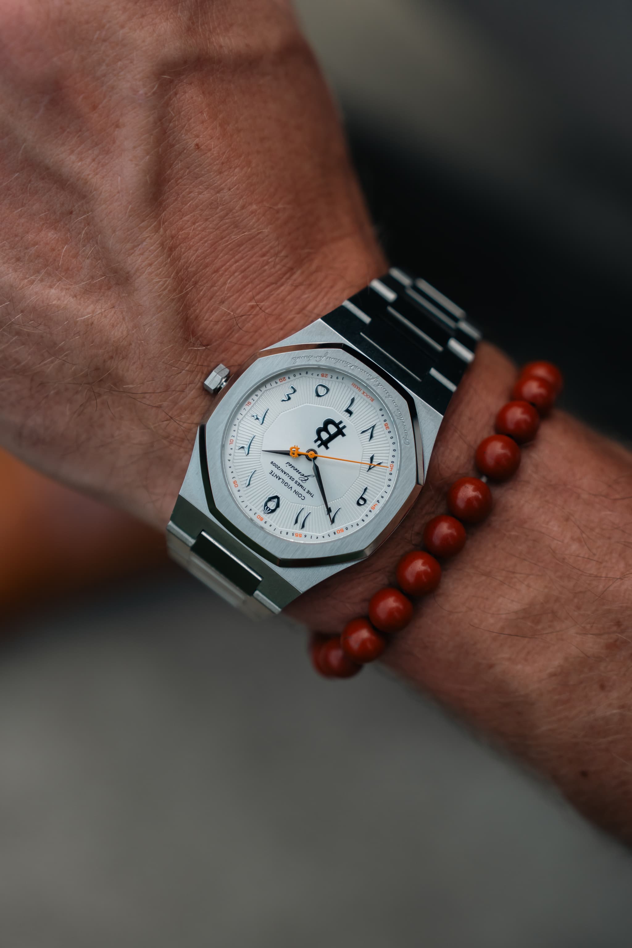 A close-up of a wrist wearing a silver-toned stainless steel watch with a white dial and black bezel, alongside a red beaded bracelet.
