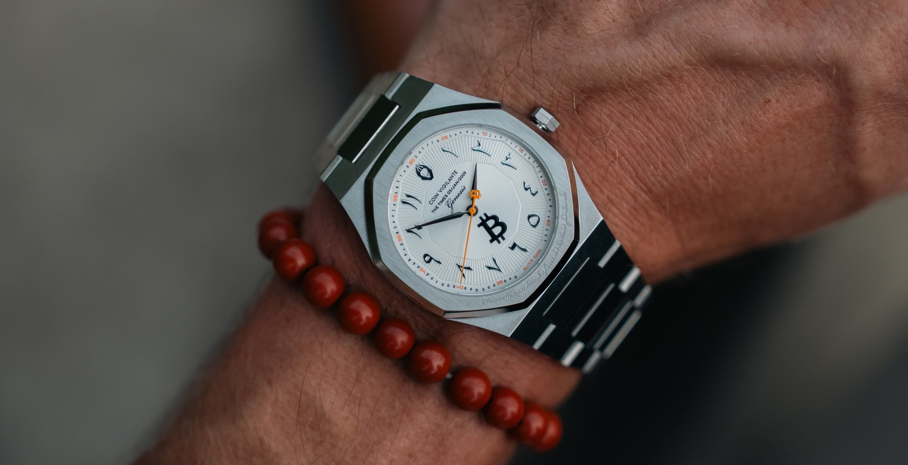 A close-up of a silver-toned wristwatch with a white dial and black octagonal case, worn on a person's wrist alongside a red beaded bracelet.