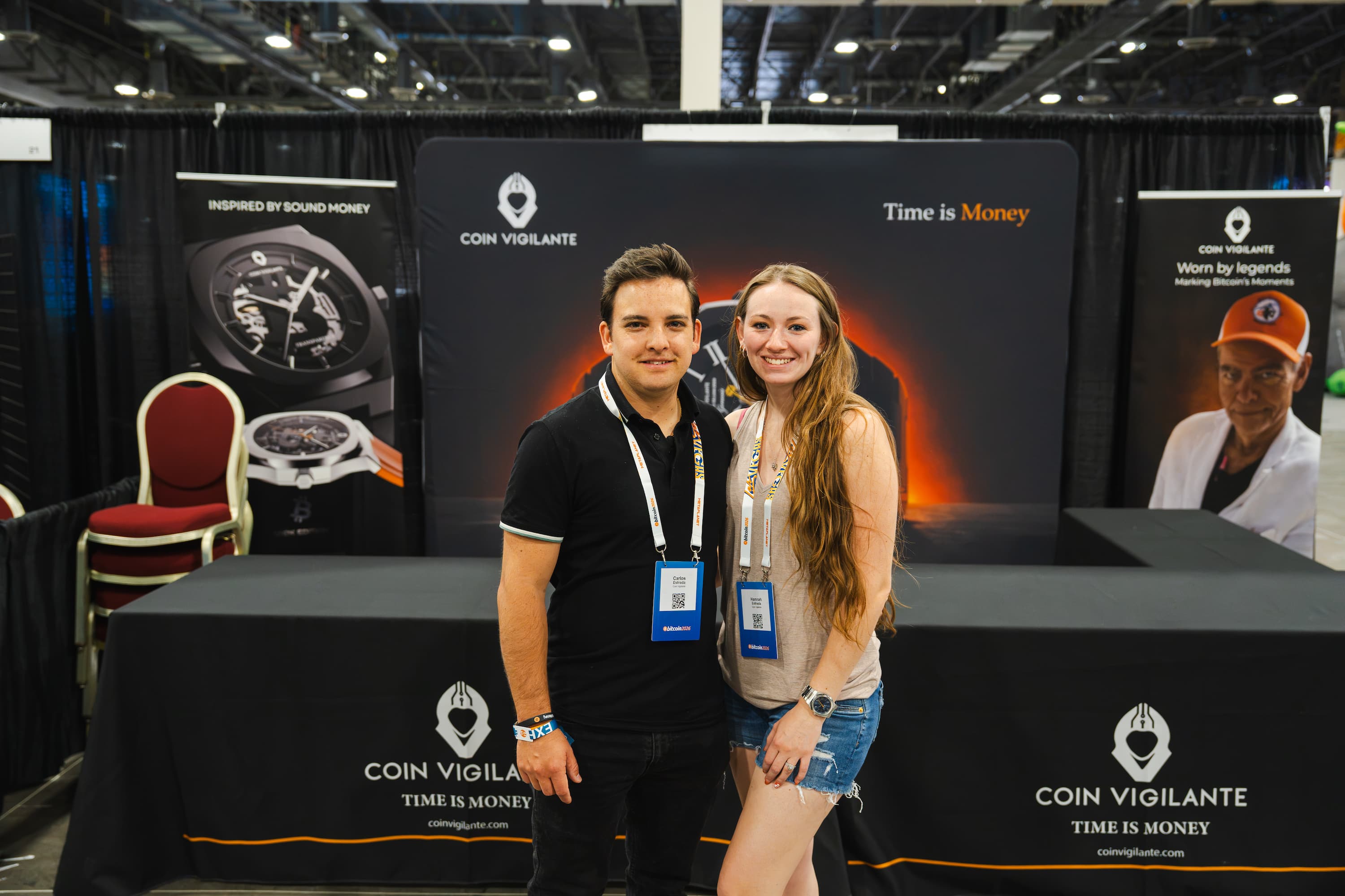 A man and a woman posing together at a Coin Vigilante booth at a trade show, with branded banners and a display table in the background.